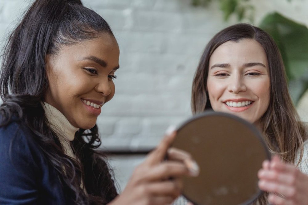 happy young female client looking in mirror and smiling near positive ethnic makeup artist