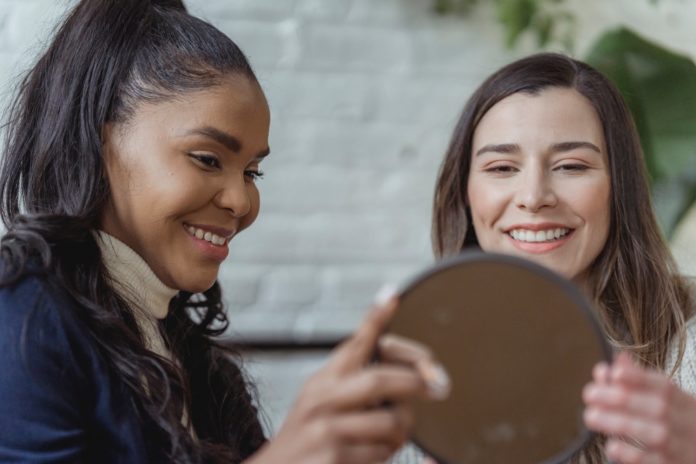 happy young female client looking in mirror and smiling near positive ethnic makeup artist
